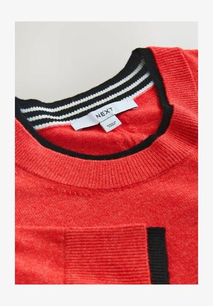 Červený svetr s žebrovaným černo-bílým proužkovaným límcem, s texturovanou tkaninou a malou kapsou na levé straně hrudníku. Viditelná značka značky.