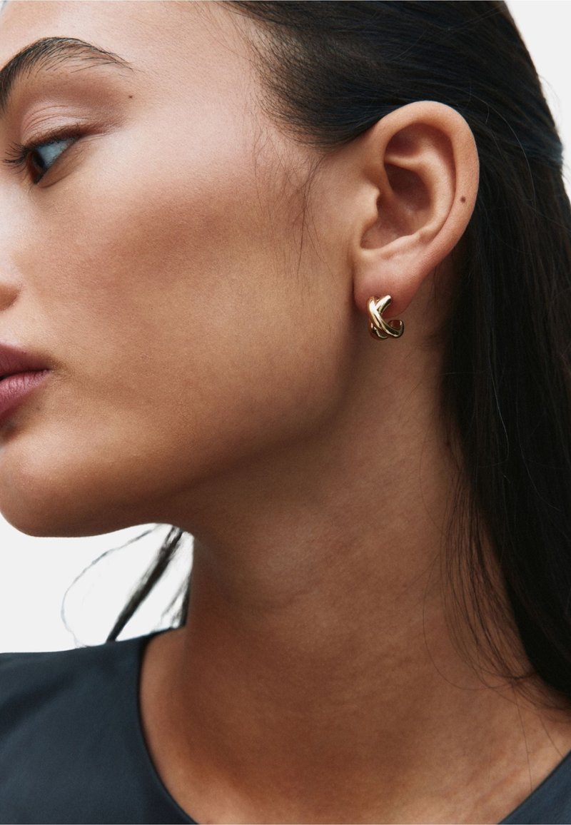 Zlata uhan obroč s prepletenim vzorcem. Gladek videz s poliranim zaključkom, nošen na levem ušesu, ki dopolnjuje temno majico.