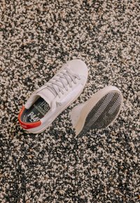 Baskets blanches avec un détail rouge au talon sur une surface texturée de gravier noir et blanc, une chaussure montrant le motif de sa semelle.