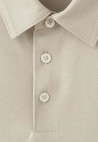 Camisa beige texturizada con cuello puntiagudo y tres botones blancos abrochados a lo largo de la tapeta frontal.