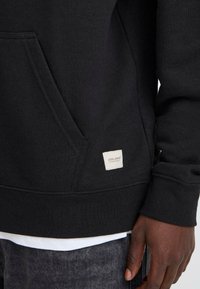 Svart hettegenser med frontlomme, ribbede mansjetter og en liten vevd etikett i kanten. Stoffet ser mykt og tykt ut.