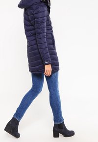 Marinblå vadderad jacka med horisontell quiltad design och huva. Bärs med åtsittande blå jeans och svarta ankelboots.