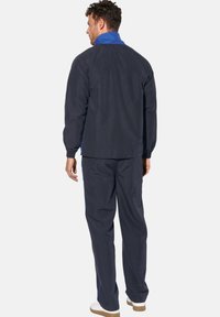 Marineblaue Oberbekleidung bestehend aus einer Jacke mit Reißverschluss und passenden Hosen. Die Jacke hat einen blauen Kragen und elastische Bündchen; die Schuhe sind hellfarben.