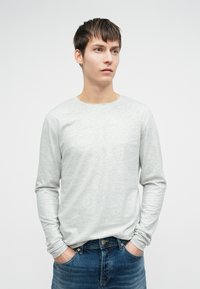 Απαλή γκρι μακρυμάνικη μπλούζα με στρογγυλή λαιμόκοψη, σε συνδυασμό με μπλε τζιν παντελόνι και χέρια που ακουμπούν στις τσέπες.