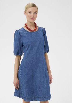 Vrouw met blond haar draagt een blauwe spijkerjurk met korte opgepofte mouwen en een ronde ketting van rode kralen, staand tegen een effen achtergrond.