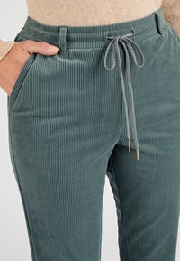 Pantaloni in corduroy verde acqua con strisce verticali, vita elastica, dettagli con coulisse e tasche laterali, che mostrano una texture morbida.