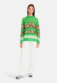 Grüner Strickpullover mit horizontalen Streifen, der floralem und Leopardenmuster zeigt, kombiniert mit weißen weitem Hosen und gemusterten Sneakers.