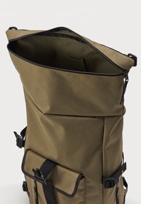 Mochila alta de color verde oliva con la cremallera superior parcialmente abierta, bolsillo frontal con correa de hebilla y correas laterales ajustables.