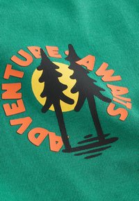 Tejido verde con un diseño gráfico. El diseño incluye un sol circular amarillo, árboles negros y texto naranja que dice "LA AVENTURA TE ESPERA".