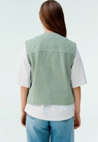 Grön ärmlös jacka, denimmaterial, något lös passform, detalj i sömmen på baksidan, lagd över en vit T-shirt och ljusblå jeans.