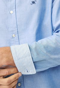 Camisa abotonada de color azul claro con una textura suave, que presenta un puño a rayas con rayas blancas y azul claro y un logotipo bordado en el pecho.