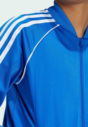 Blå atletisk jakke med hvide striber på skulderne, lynlås lukning og ribbet krave. Glat, letvægtsstof med et sporty design.