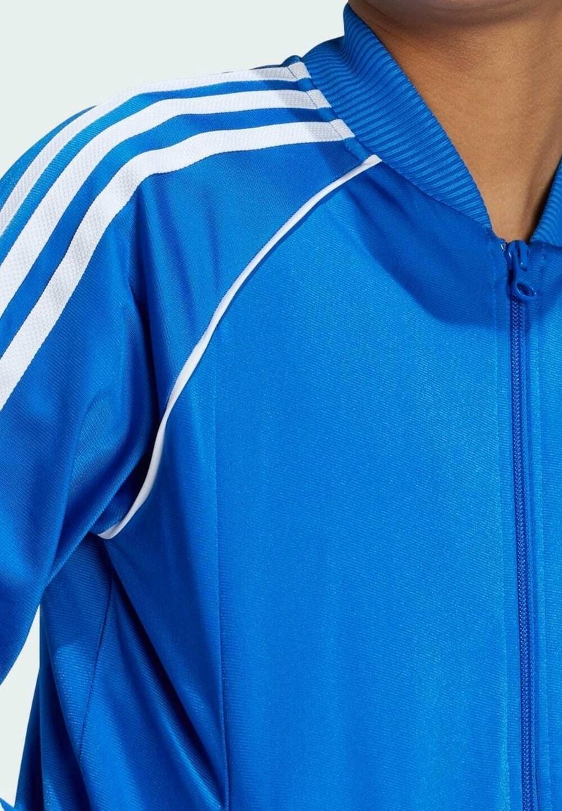 Modrá sportovní bunda s bílými pruhy na ramenou, s zipovým zapínáním a žebrovaným límcem. Hladká, lehká tkanina se sportovním designem.