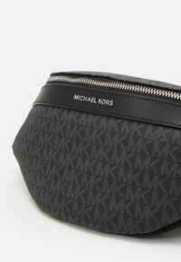 Marsupio in tessuto nero con motivo 'MK' in rilievo, una tracolla in pelle liscia e una cerniera in metallo argentato con etichetta personalizzata.
