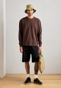 Brown long-sleeve shirt, black shorts, black sneakers with gum soles, holding a beige bag, wearing an olive bucket hat. Wood and neutral background.