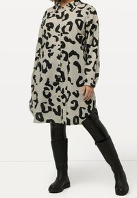 Knoopstrijkl shirtjurk in beige met een zwart luipaardpatroon. Lange mouwen, kraag en zijsplitten. Gecombineerd met zwarte kniehoge laarzen.