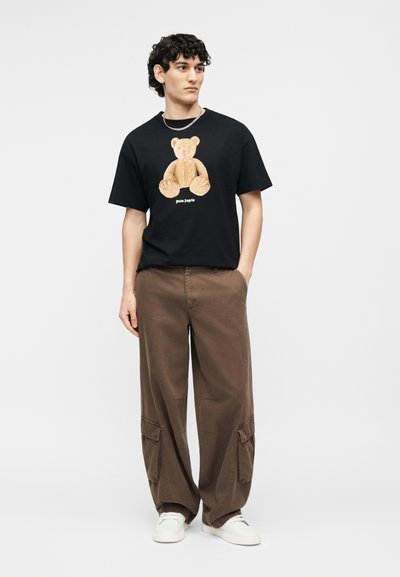 Junger Mann mit lockigem Haar, der ein schwarzes T-Shirt mit Teddy-Print, braune Cargo-Hosen, weiße Turnschuhe und eine silberne Kettenhalskette trägt.