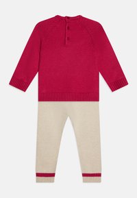 Maglione rosso a maglia con scollatura arrotondata e tre bottoni sul retro, abbinato a leggings crema con polsini a righe rosse.