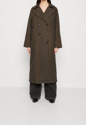 Kvinde iført en lang, mørkebrun dobbeltspændt trenchcoat med brede revers, kombineret med sorte bukser med vide ben og sorte sko.