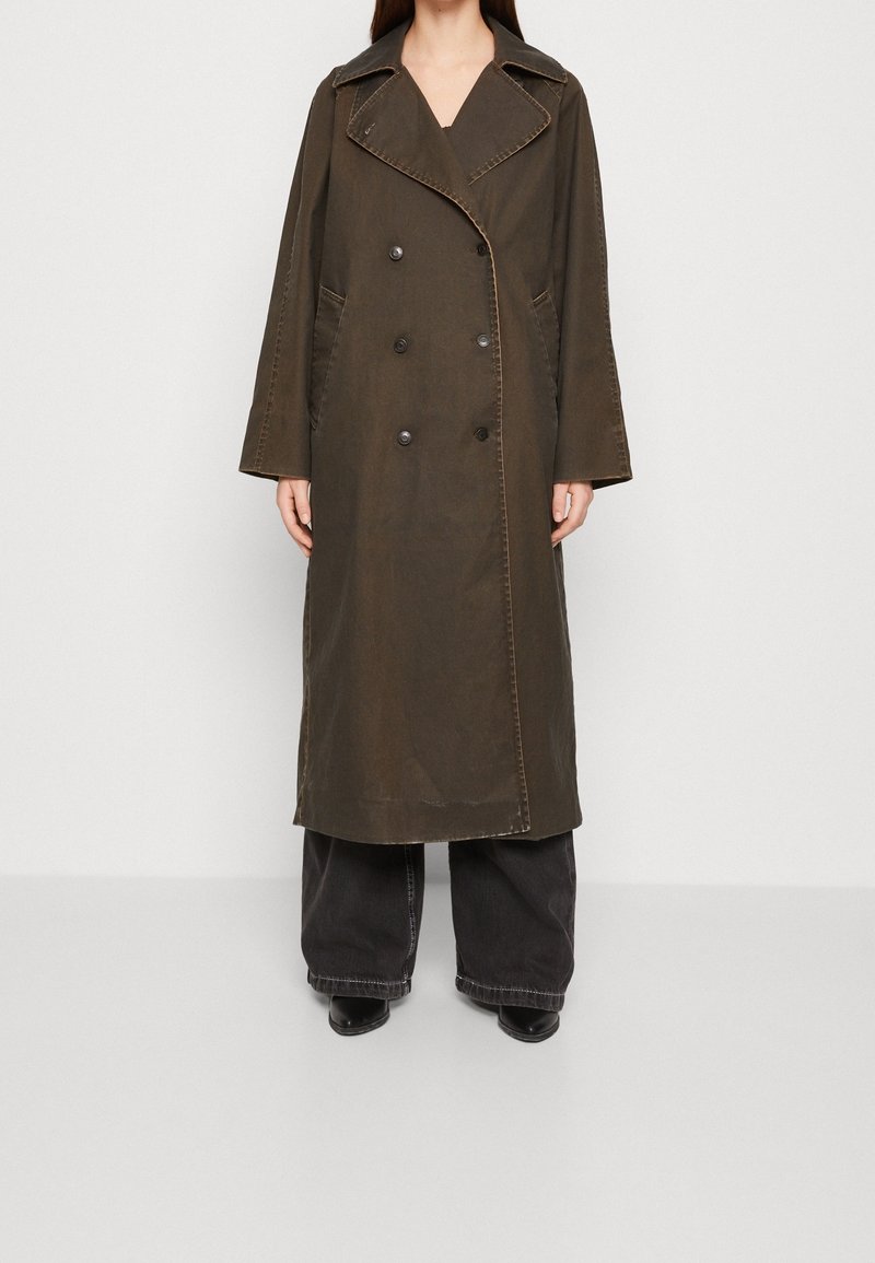 Femme portant un long trench-coat marron foncé à double boutonnage avec de larges revers, associé à un pantalon noir à jambes larges et des chaussures noires.