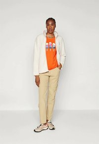 Giacca in pile bianca, t-shirt arancione con design grafico di ghiacciolo, pantaloni beige e sneakers chiare. Modello in piedi davanti a uno sfondo neutro.