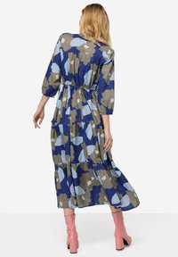 Vestido midi floral con un patrón en azul, gris y crema, cintura fruncida, mangas largas y falda en capas; combinado con botas de tacón rosas.