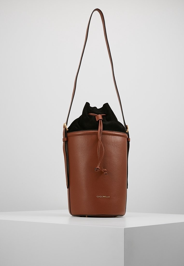 Borsa secchiello in pelle marrone con chiusura in tessuto nera a cordoncino e tracolla lunga, posizionata su un piedistallo bianco.