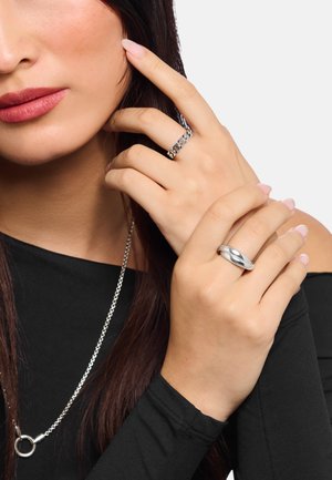 Woman wearing a black top with two silver rings on fingers and a silver chain necklace, touching her face with one hand.