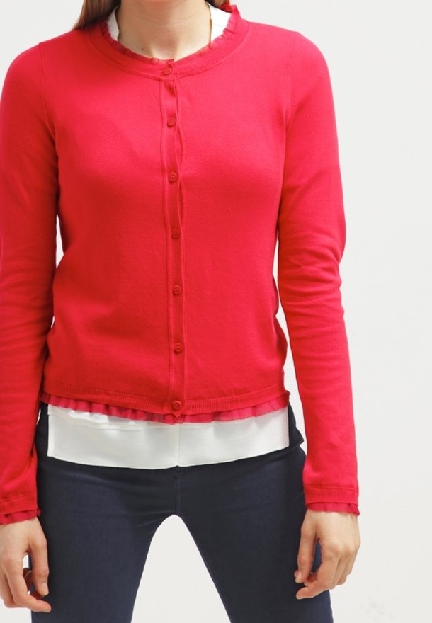Femme portant un cardigan rouge vif boutonné sur un chemisier blanc et un pantalon sombre, debout devant un fond clair uni.