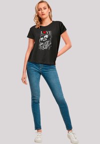 Black T-shirt featuring skeleton design with red heart and text "LOVE IS ALL YOU NEED," paired with blue skinny jeans and white sneakers.
