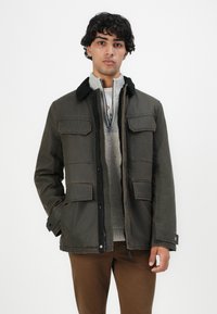 Giovane uomo con capelli neri e ricci, indossa una giacca verde scuro sopra un maglione grigio a zip e pantaloni marroni, in piedi contro uno sfondo bianco.