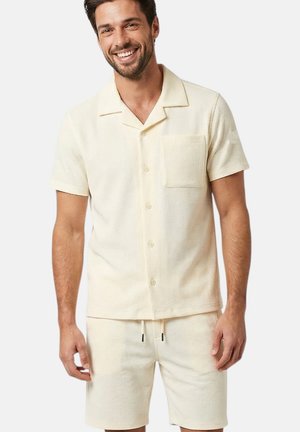 Homme souriant portant une chemise boutonnée texturée crème à manches courtes avec poche poitrine et un short assorti à cordon de serrage.