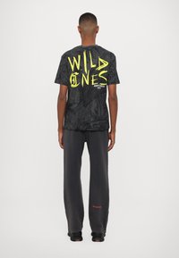 T-shirt noir avec le texte "WILD ONES" en jaune fluo, présentant un motif de plante texturé. Associé à un pantalon gris foncé ample.