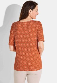 Korte mouwen top in oranje met een geometrisch crèmekleurig patroon. Heeft een ronde halslijn en een relaxed fit. Stof lijkt glad te zijn.