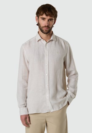Uomo che indossa una camicia a righe crema e beige a maniche lunghe con bottoni e pantaloni beige su uno sfondo grigio chiaro uniforme.