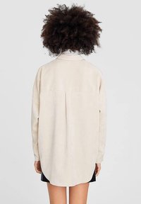 Camicia oversize color crema con maniche lunghe, colletto a bottoni, dettaglio della piega sul retro e spacchi laterali, realizzata in un tessuto leggero e morbido.