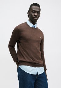 Barna kötött pulóver V-nyakú kialakítással, világoskék galléros ing felett viselve. A ruhát farmernadrág egészíti ki.