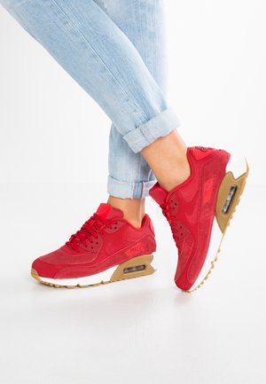 Benedenbenen in lichtblauwe opgerolde spijkerbroeken met rode Nike Air Max sneakers met witte tussenzolen en bruine zolen op een witte ondergrond.