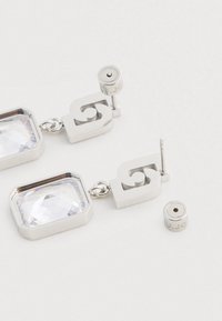 Sudraba pilienu auskari ar lieliem caurspīdīgiem taisnstūrveida kristāliem un ģeometriskām izgriezumu detaļām, attēloti ar noņemamiem aizmugurējiem stiprinājumiem uz baltas virsmas.