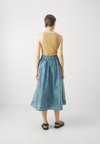 Top in cotone rigato marrone, gonna maxi blu trasparente con vita elastica e sandali neri con decorazioni in metallo. Sfondo semplice.