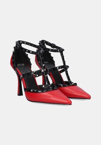 Zapatos de tacón alto de cuero rojo con tiras de charol negro que presentan tachuelas en forma de pirámide. Diseño de punta afilada y tacón de estilete. Apariencia elegante y audaz.