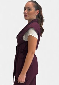 Gilet senza maniche color bordeaux con design da blazer, caratterizzato da spalle larghe, bottoni e vita con cintura, indossato su una camicia bianca. Tessuto strutturato.