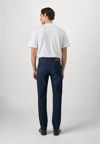 Jeans de mezclilla azul oscuro con corte recto, que cuentan con bolsillos traseros y un parche de piel marrón en la parte posterior, combinados con una camiseta blanca lisa.