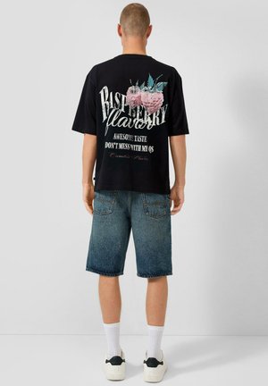 Person trägt ein schwarzes T-Shirt mit großem Himbeer-Design und dem Text "Raspberry Flavor, Awesome Taste, Don't Mess With My Qs" auf dem Rücken, dazu Jeansshorts.