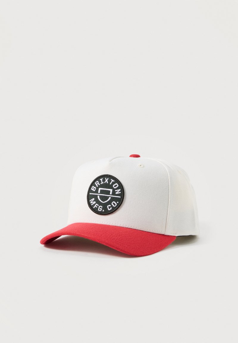 Λευκό και κόκκινο καπέλο μπέιζμπολ με μαύρο κυκλικό έμβλημα που γράφει "BRIXTON MFG. CO." στην μπροστινή πλευρά, σε απλό φωτεινό φόντο.