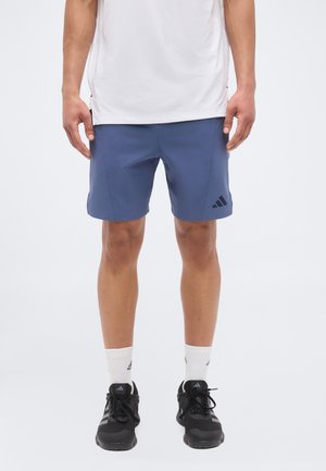 Mężczyzna ubrany w niebieskie szorty Adidas, białe skarpetki Adidas, czarne buty do biegania Adidas i białą koszulkę, stoi prosto na jednolitym tle.