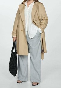 Manteau beige à double boutonnage sur une chemise blanche. Pantalons amples à rayures gris. Sac fourre-tout tissé noir et sandales noires.