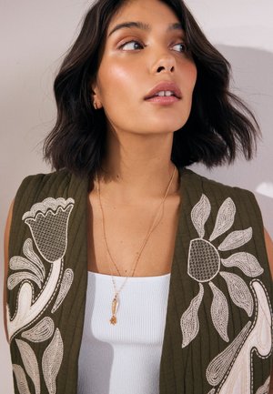 Young woman with dark wavy hair wears olive green vest with large beige floral embroidery over white tank top, looking to the side.