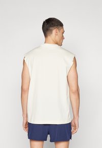 Sleeveless cream cotton shirt with a round neckline, featuring raw edges on shoulders, paired with navy shorts.