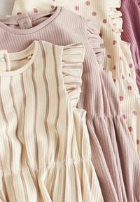 Tri otroške obleke: ena kremna s subtilnimi črtami in volančki, ena svetlo roza z volančki in ena temno roza s pikami.